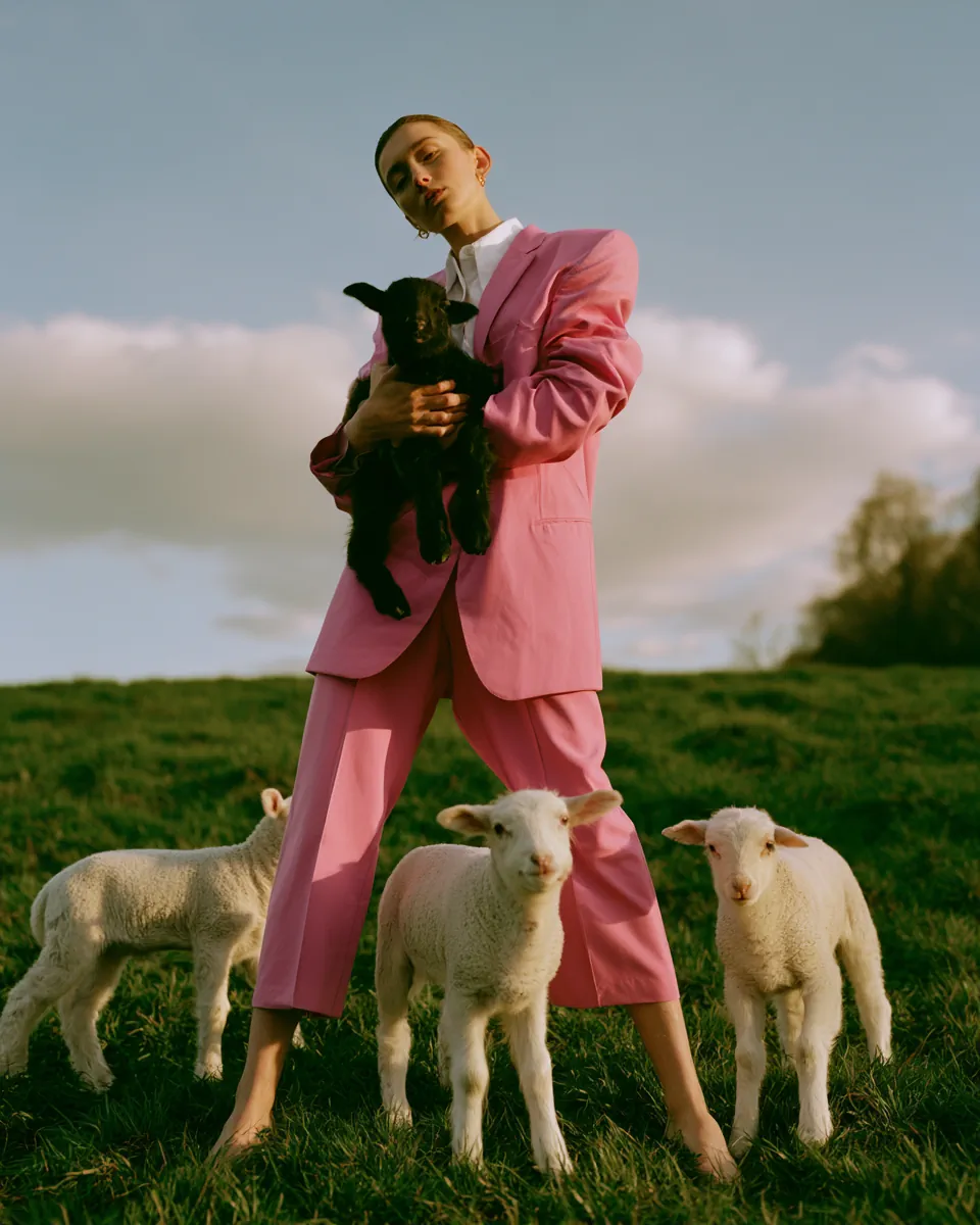 Young European woman, light skin, relaxed expression, wearing a vivid pink blazer over a white shirt, standing full height on a lush green meadow, cradling a small black lamb in both arms. Three white lambs roam around his feet. Dynamic low angle perspective looking slightly upward, full figure against open sky with soft clouds. Golden hour sunlight from the side casting warm directional shadows, rim lighting the pink blazer. Playful, editorial, fashion meets pastoral mood. Shot on Hasselblad X2D 100C, 35mm f/2.0 wide angle, Kodak Portra 400 film aesthetic, fine art environmental portrait, natural soft skin tones, ultra sharp clothing and animal texture detail, shallow background depth of field, muted warm green and soft pink palette with creamy highlights, gentle film grain, minimal retouching, professional editorial quality.